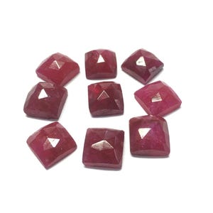 May include: Ten faceted ruby gemstones in a square shape. The gemstones are a deep red color and have a polished finish.