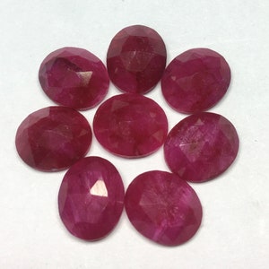 Op de afbeelding: Tien facetgeslepen ovale robijn edelstenen in verschillende tinten roze en rood. De edelstenen zijn in een cirkelvormige opstelling op een witte achtergrond geplaatst.