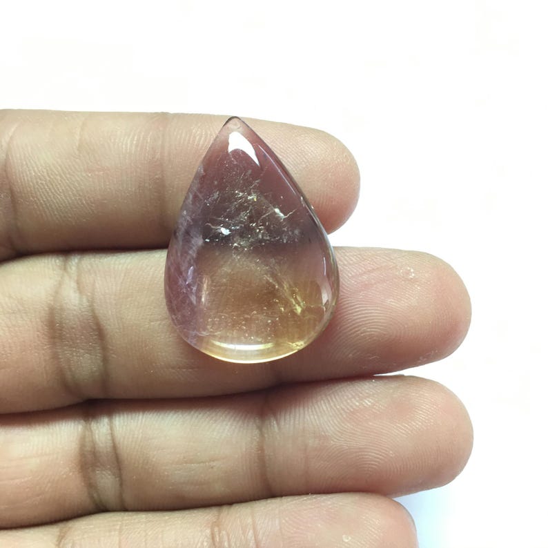 Puede incluir: Una piedra preciosa cabuj&oacute;n en forma de l&aacute;grima con un degradado de rosa a amarillo. La piedra preciosa es transl&uacute;cida y tiene una superficie lisa y pulida.