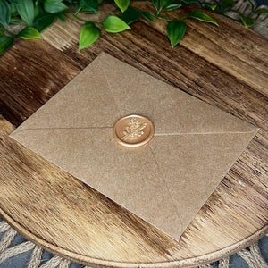 Puede incluir: Un sobre de papel marr&oacute;n con un sello de lacre dorado con un dise&ntilde;o floral.