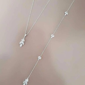 May include: A silver necklace with a delicate pendant and a longer chain with multiple small pendants. Each pendant features a cluster of small, clear stones in a leaf shape.