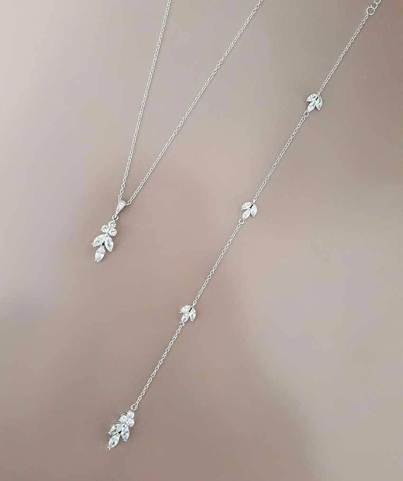 May include: A silver necklace with a delicate pendant and a longer chain with multiple small pendants. Each pendant features a cluster of small, clear stones in a leaf shape.