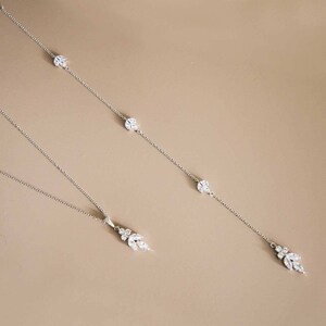 May include: Two silver necklaces with crystal accents. The shorter necklace has a cluster of crystals at the end. The longer necklace has three crystal accents and a cluster of crystals at the end.