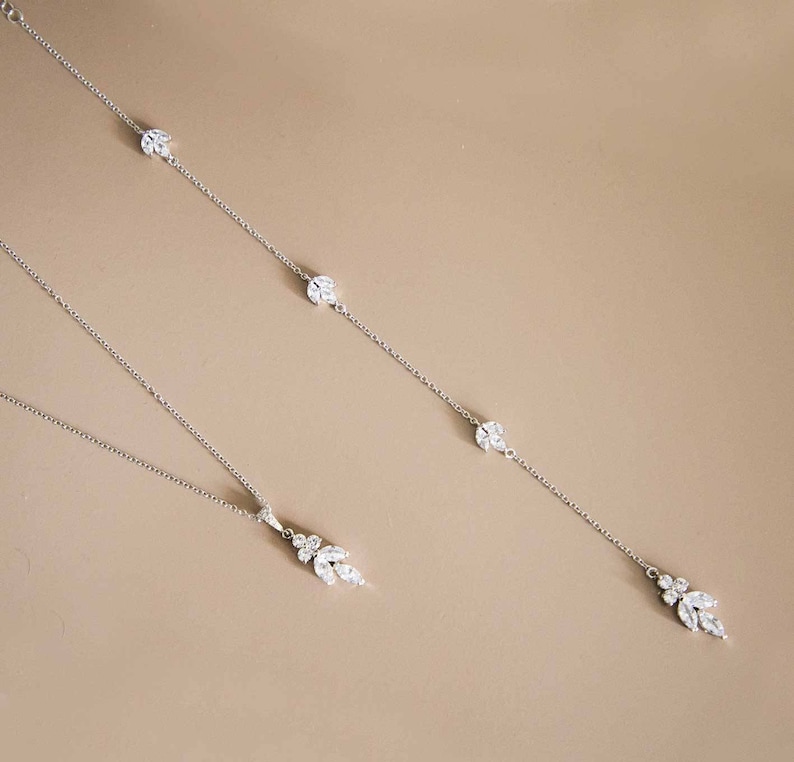May include: Two silver necklaces with crystal accents. The shorter necklace has a cluster of crystals at the end. The longer necklace has three crystal accents and a cluster of crystals at the end.