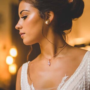 May include: A woman wearing a white wedding dress with a delicate rose gold necklace featuring a small floral pendant. She has her hair pulled back in a bun and is wearing pearl earrings.