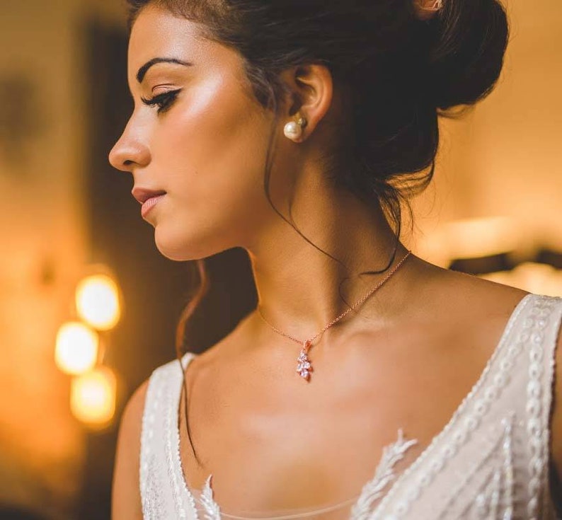May include: A woman wearing a white wedding dress with a delicate rose gold necklace featuring a small floral pendant. She has her hair pulled back in a bun and is wearing pearl earrings.