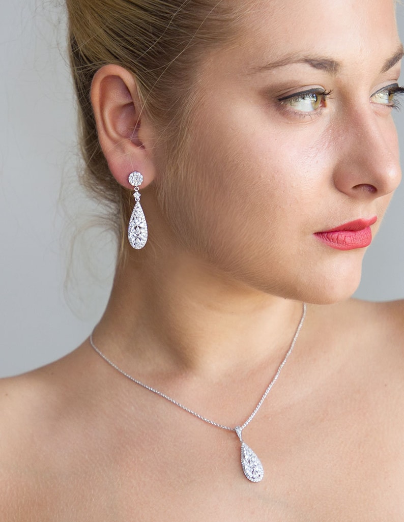 May include: A silver necklace and earrings set with a teardrop-shaped pendant and matching earrings, both adorned with sparkling clear stones.