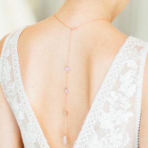 May include: A rose gold back necklace with a delicate chain and small crystal accents. The necklace is worn by a person with a white lace dress.