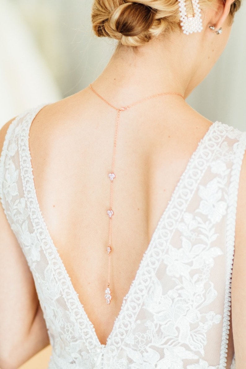 May include: A rose gold back necklace with a delicate chain and small crystal accents. The necklace is worn by a person with a white lace dress.