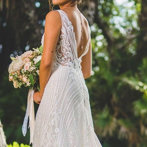 May include: A bride wearing a white lace wedding dress with a low back and a delicate beaded necklace. She is holding a bouquet of pink and white flowers.