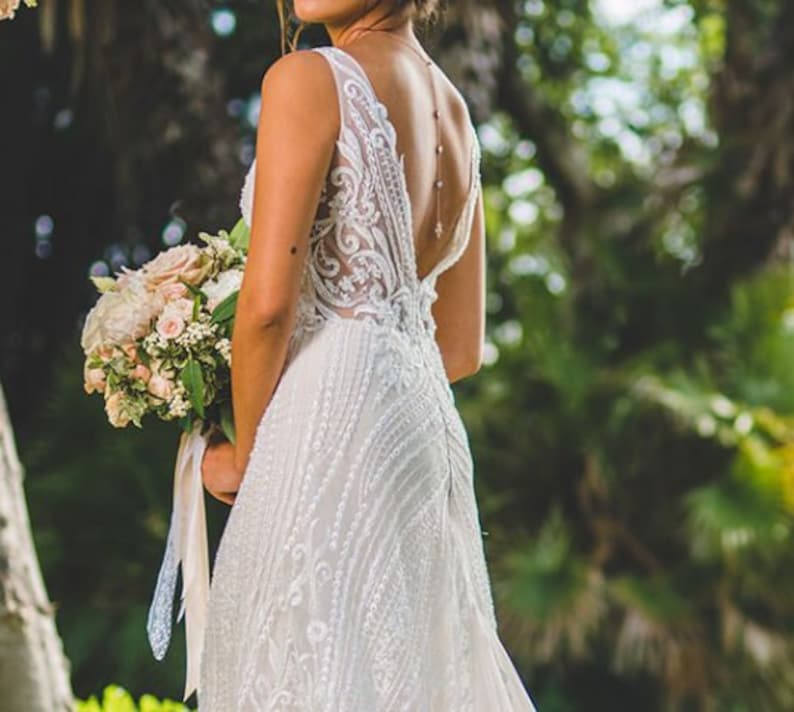 May include: A bride wearing a white lace wedding dress with a low back and a delicate beaded necklace. She is holding a bouquet of pink and white flowers.