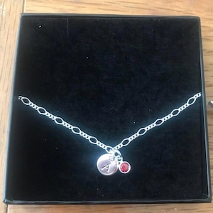 May include: A silver chain necklace with a silver charm featuring the letter 'A' and a small red gemstone.