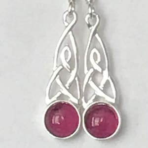 May include: A pair of silver drop earrings. Each earring has a Celtic knot design and a round, deep pink stone at the base. The earrings have a simple hook closure.