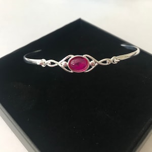 May include: A silver bangle bracelet featuring a pink oval-shaped gemstone set in the centre. The bracelet has a delicate, intricate design.