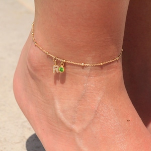 May include: Gold anklet with a small green gemstone and a charm with the letter "R" set with small clear stones.