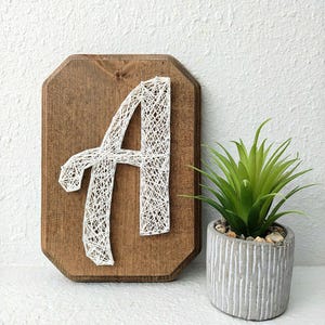 May include: White string art of the letter A on a wooden plaque with a rustic finish. The plaque is mounted on a white wall next to a small potted succulent plant.