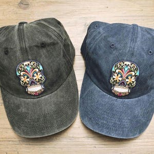 May include: Two baseball caps, one in charcoal gray and one in faded navy blue, both featuring a colorful embroidered sugar skull patch on the front panel. Each cap has a six-panel construction with a curved brim and matching top button.