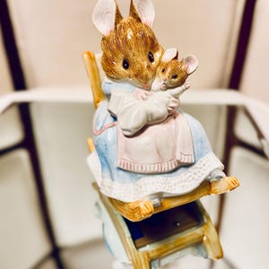 Puede incluir: Figura de cerámica de una madre ratón sosteniendo a un bebé ratón. Los ratones son marrones y blancos, visten un vestido azul y rosa, y están sentados en una mecedora de madera. Diseño detallado.