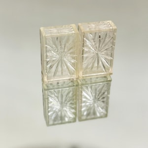 May include: Four clear, rectangular glass boxes are stacked in two pairs. Each box features a starburst design on the front and back, with vertical lines on the sides. The boxes are reflected on a light-colored surface.