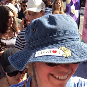 May include: A woman wearing a blue denim bucket hat with a red heart and the text "I &hearts; Paris" embroidered on the front. The hat has a yellow croissant embroidered on the right side.