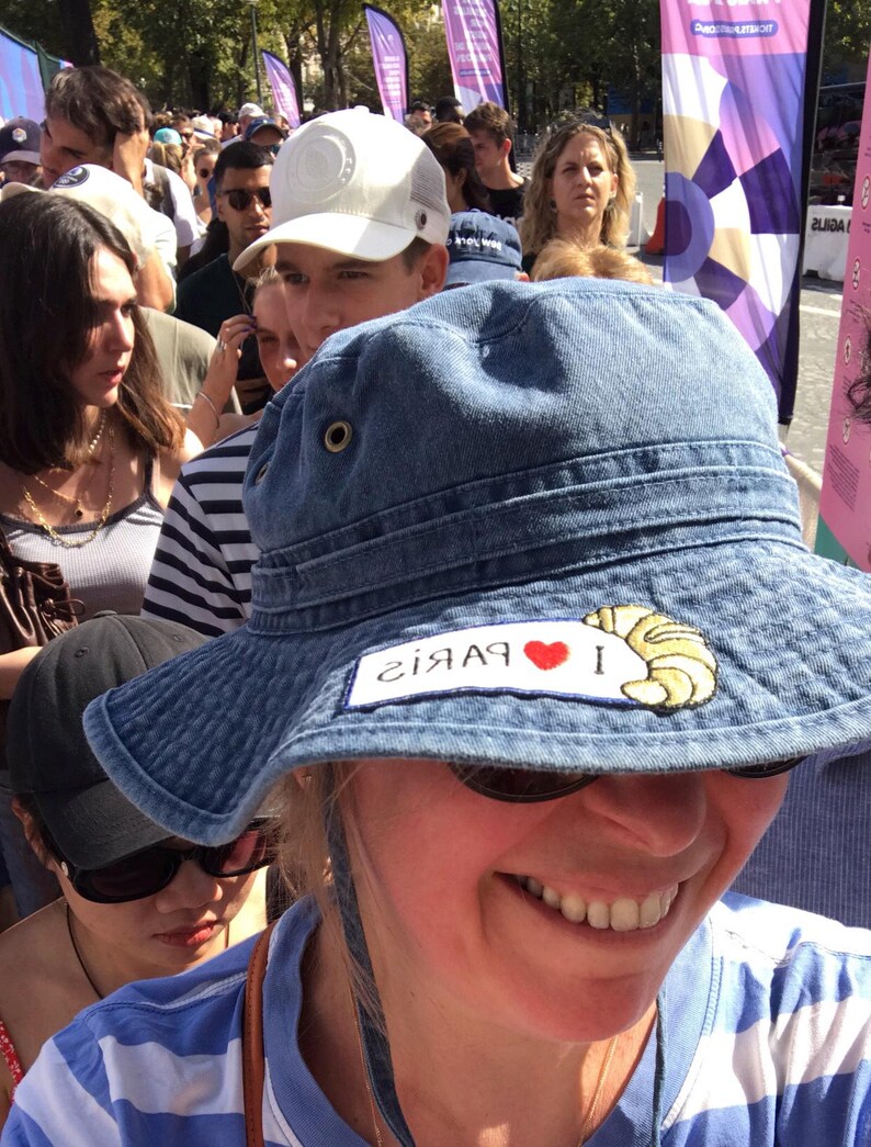 May include: A woman wearing a blue denim bucket hat with a red heart and the text "I &hearts; Paris" embroidered on the front. The hat has a yellow croissant embroidered on the right side.