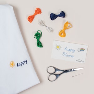 Peut inclure: Un tissu blanc avec les mots "happy" et un soleil brodés dessus. Il y a cinq bobines de fil à broder de différentes couleurs, une paire de ciseaux, une aiguille et une carte avec les mots "happy mama" et un arc-en-ciel.