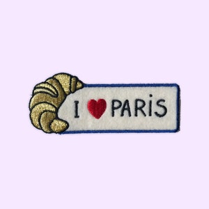 May include: Embroidered patch with a gold croissant and a white banner with blue trim. The banner says "I love Paris" with a red heart.