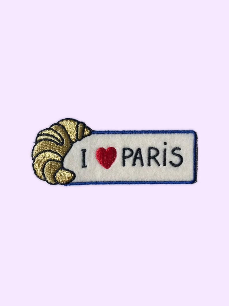 May include: Embroidered patch with a gold croissant and a white banner with blue trim. The banner says "I love Paris" with a red heart.