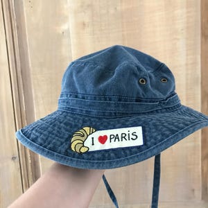 May include: A blue denim bucket hat with a patch that says "I love Paris" and a yellow croissant graphic.