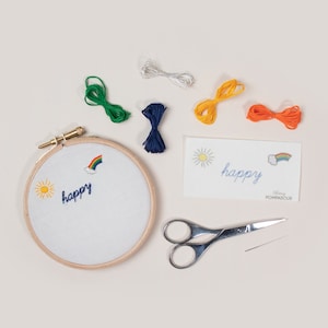 May include: A white embroidery hoop with a rainbow, sun, and the word "happy" embroidered on it. The hoop is surrounded by embroidery floss in green, blue, yellow, and orange, a silver thread, a pair of scissors, and a needle.