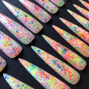 May include: A set of long, white, stiletto-shaped false nails with a colorful confetti design. The nails are arranged in rows on a black surface.