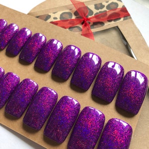 May include: Set of 10 purple glitter press-on nails with a holographic finish. The nails are arranged in two rows on a brown surface.