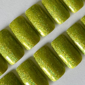 May include: Close-up of several lime green glitter press-on nails. The nails are rectangular with rounded edges and are arranged diagonally. The glitter is evenly distributed across the surface, creating a sparkling effect.