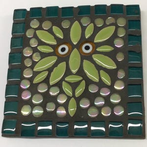 Puede incluir: Azulejo de mosaico cuadrado con diseño de búho. Los ojos del búho son blancos con centros marrones, rodeados de azulejos en forma de hoja verde claro. El fondo es gris oscuro con azulejos redondos iridiscentes y azulejos cuadrados turquesa en el borde.