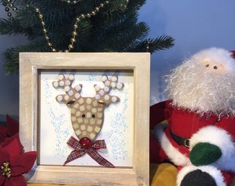 May include: A framed Christmas decoration featuring a reindeer mosaic with a red plaid bow. The reindeer has a brown mosaic face and antlers with white and gold accents. A small Christmas tree and Santa Claus doll are in the background.