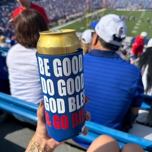 May include: A blue beverage insulator with white text that reads "BE GOOD DO GOOD GOD BLESS & GO BILLS" holds a gold can. The image is taken at a stadium with a crowd in the background.
