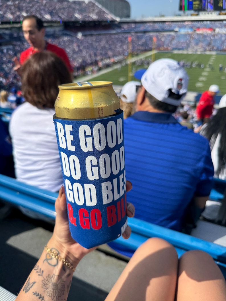 May include: A blue beverage insulator with white text that reads "BE GOOD DO GOOD GOD BLESS & GO BILLS" holds a gold can. The image is taken at a stadium with a crowd in the background.