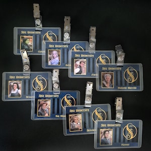 May include: A collection of clear plastic name badges with metal clips. Each badge features a portrait, the words "Shiz University", and a gold and blue design. Names include Elphaba, Nessarose, and Madame Morrible.