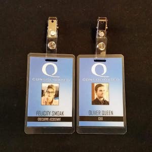 May include: Two blue and white plastic ID badges with silver clips. The top badge reads "Q Consolidated" and "Oliver Queen, CEO". The bottom badge reads "Q Consolidated" and "Felicity Smoak, Executive Assistant".