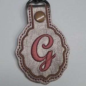 May include: A metallic silver keychain fob with a scalloped edge, featuring a large embroidered pink letter G in the center. The fob is secured with a bronze-colored snap button and attached to a black metal lobster claw clasp for easy attachment.