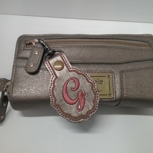 May include: A metallic pewter-colored rectangular wallet with a zippered top and a gold-toned metal brand plate. Attached to the zipper pull is a scalloped, metallic pewter-colored leatherette keychain featuring an embroidered pink letter G.