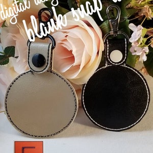 May include: Two blank snap tabs, one tan and one black, with metal clasps. The tan tab has black stitching, and the black tab has white stitching. The text "digital design file blank snap tab" is at the top. The logo "EmbroideryZzz" is at the bottom.