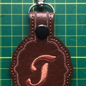 May include: A brown faux leather keychain with a scalloped edge and a silver metal lobster clasp. The keychain features a black snap button and an embroidered copper-colored letter T in the center, with visible stitching around the perimeter.