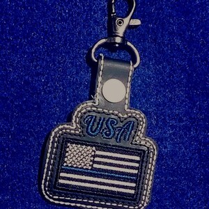 Puede incluir: Un llavero con un mosquet&oacute;n de metal y un colgante de polipiel. El colgante presenta la bandera de EE. UU. con una franja azul y la palabra "USA" en escritura azul. Los bordes est&aacute;n cosidos con hilo blanco.