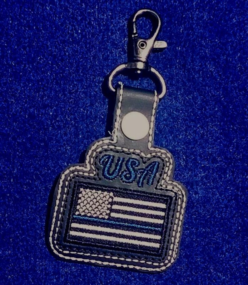 Puede incluir: Un llavero con un mosquet&oacute;n de metal y un colgante de polipiel. El colgante presenta la bandera de EE. UU. con una franja azul y la palabra "USA" en escritura azul. Los bordes est&aacute;n cosidos con hilo blanco.