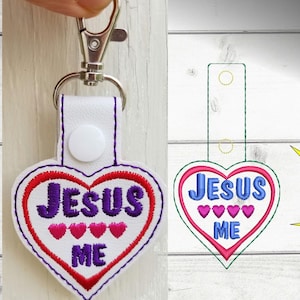 Puede incluir: Llavero de cuero blanco en forma de corazón con bordados rojos y morados. El llavero presenta el texto "JESUS ME" con tres corazones rosas entre las palabras. El llavero tiene un cierre de metal plateado.