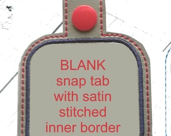 Puede incluir: Una trabilla de cierre a presión de cuero gris con un botón rojo y un borde interior cosido en satén. El texto "BLANK snap tab with satin stitched inner border" está impreso en rojo en la trabilla.
