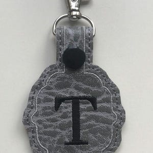 May include: A gray keychain featuring a textured, pebbled surface with a scalloped edge. The center displays a large, embroidered black letter T. A black snap button secures the top loop, which is attached to a silver-toned metal lobster clasp.