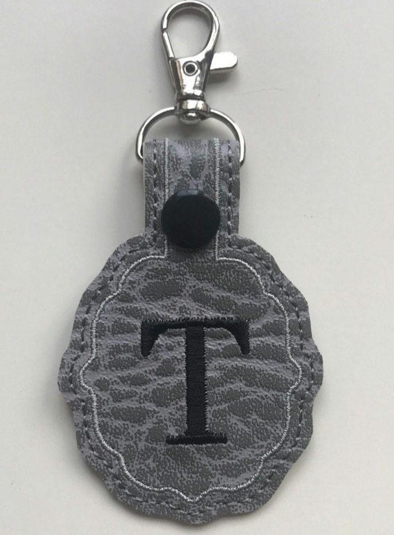 May include: A gray keychain featuring a textured, pebbled surface with a scalloped edge. The center displays a large, embroidered black letter T. A black snap button secures the top loop, which is attached to a silver-toned metal lobster clasp.