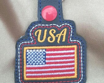 May include: A navy blue leather keychain with a red button and a silver ring. The keychain features the word "USA" in gold lettering and an embroidered American flag with red, white, and blue details.